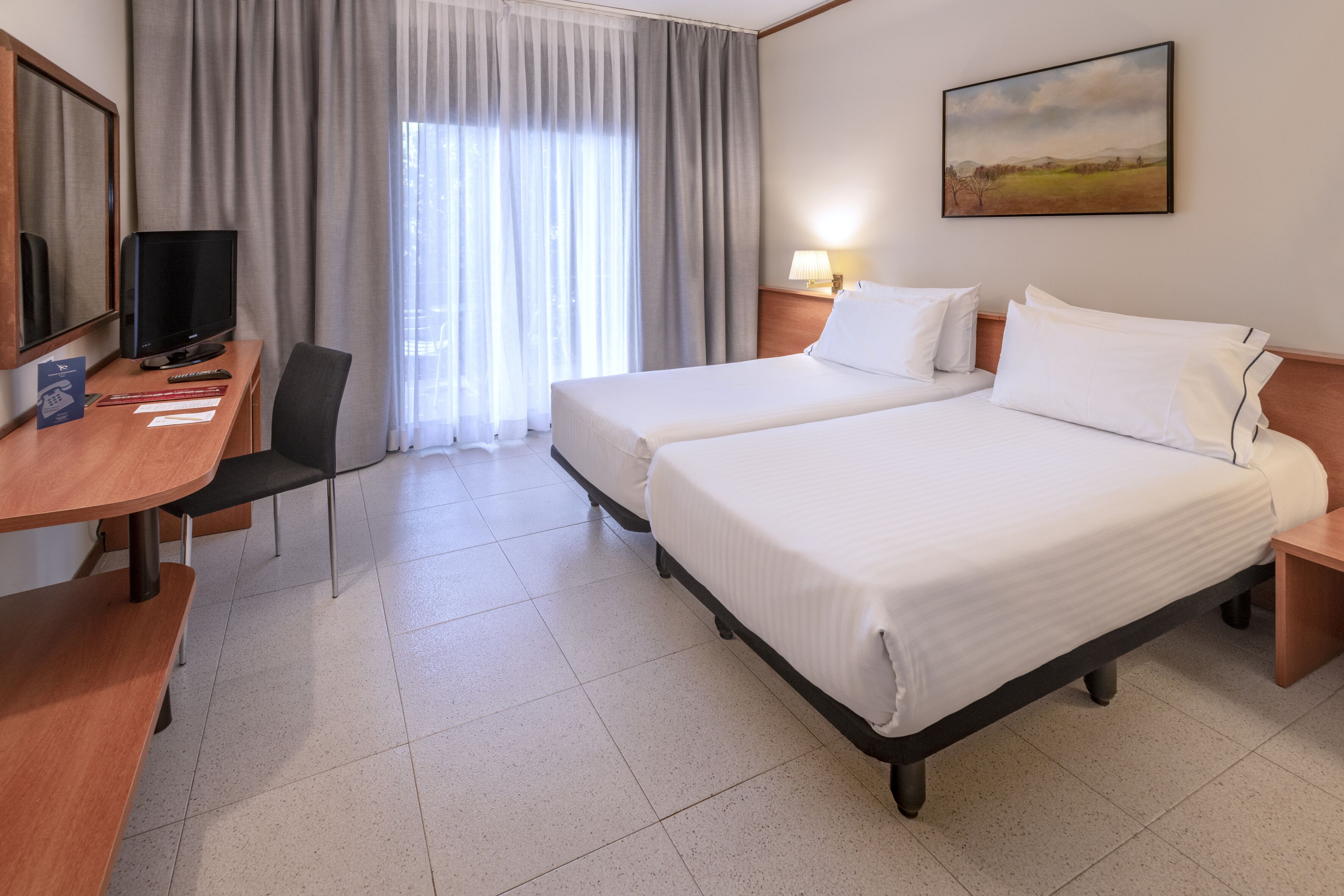 A hotel room with two beds, a desk with a TV, a chair, and a window with curtains.