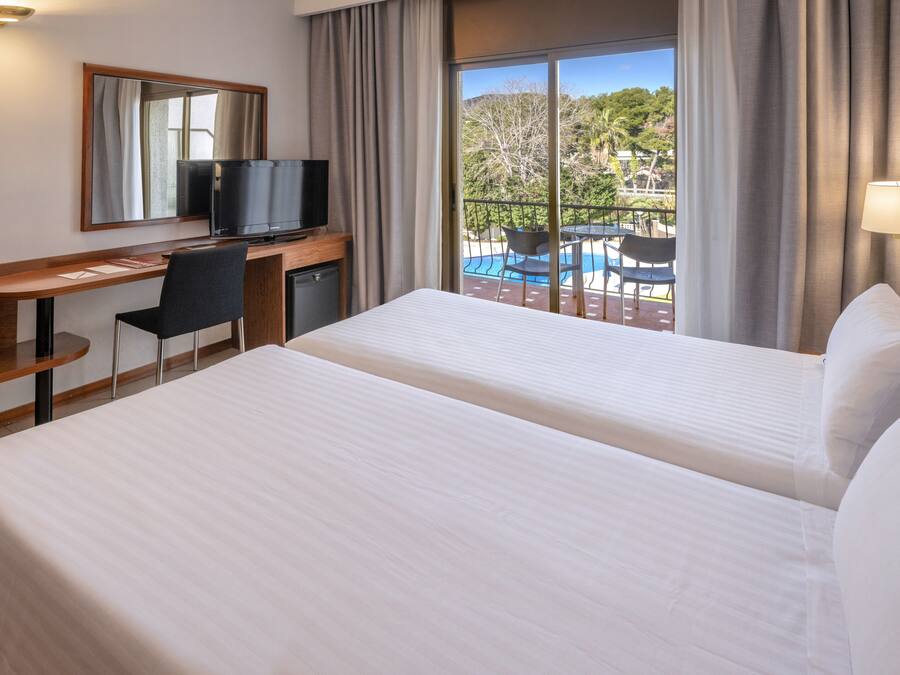 Habitación de hotel con dos camas, un escritorio con televisor, una silla y vistas a una piscina y árboles.