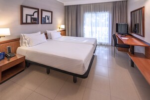 A hotel room with two beds, a desk, a chair, a TV, and a large window with curtains.