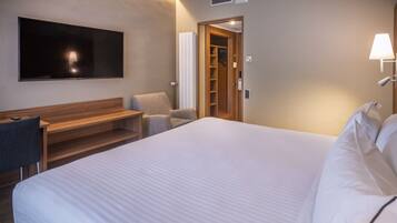 Quarto de hotel com uma cama grande, uma escrivaninha, uma cadeira, uma televisão, um guarda-roupa e uma luminária.