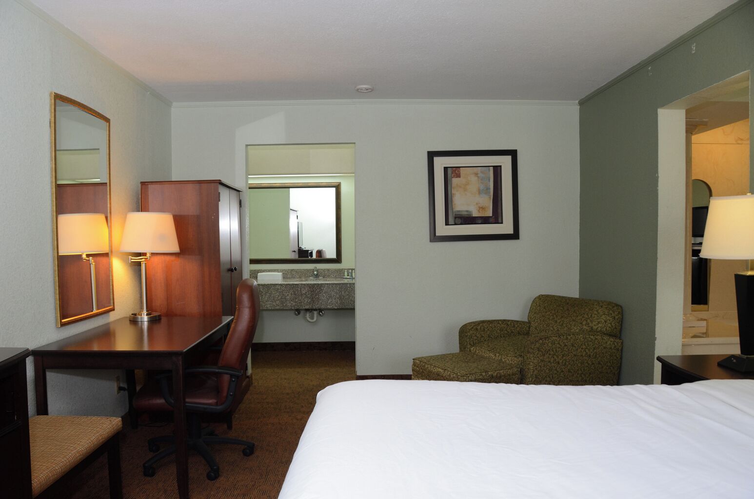 Habitación de hotel con cama, escritorio con silla, espejo, un cuadro en la pared y baño visible a través de una puerta abierta.