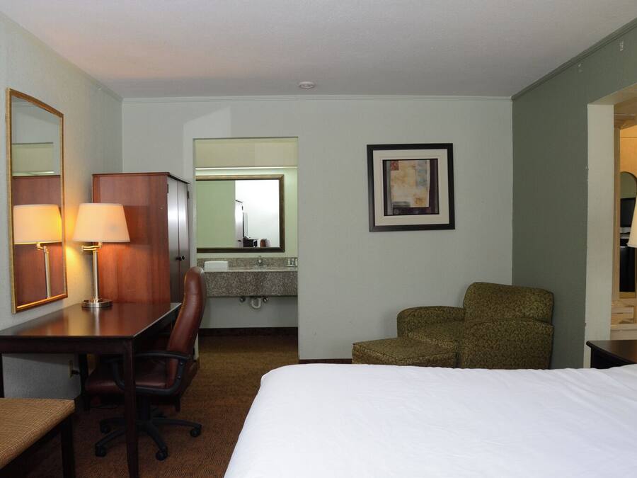 Habitación de hotel con cama, escritorio con silla, espejo, un cuadro en la pared y baño visible a través de una puerta abierta.