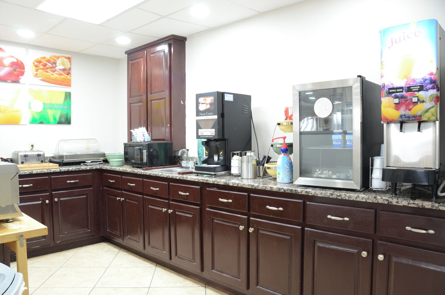 A kitchen with dark wooden cabinets, a coffee machine, a refrigerator, and a juice dispenser.