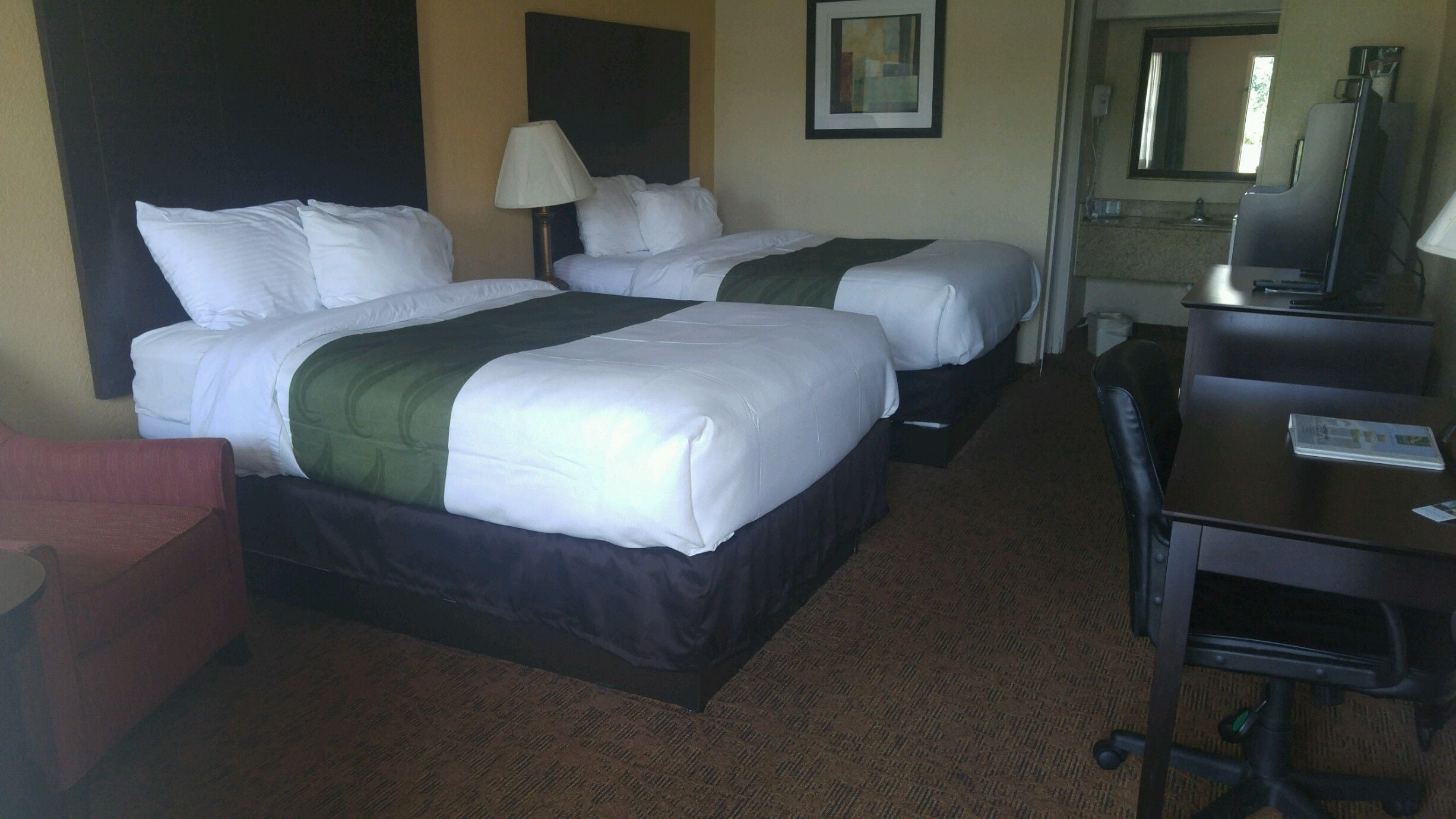 A hotel room with two beds, a desk, a chair, a lamp, and a mirror.