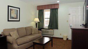 A living room with a beige sofa, a small table, a lamp, a window with curtains, an air conditioning unit, and a door. - Quality Inn & Suites Covington (Covington)