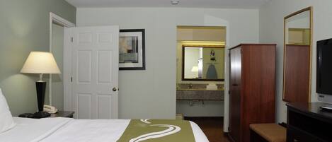 A hotel room with a bed, a desk with a lamp, a television, a wardrobe, and a bathroom visible through an open door.