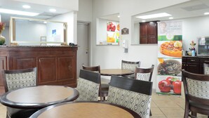 A breakfast area with wooden tables and chairs, a counter with a phone, and a wall with food images and a sign inviting people to join for breakfast. - Quality Inn & Suites Covington (Covington)