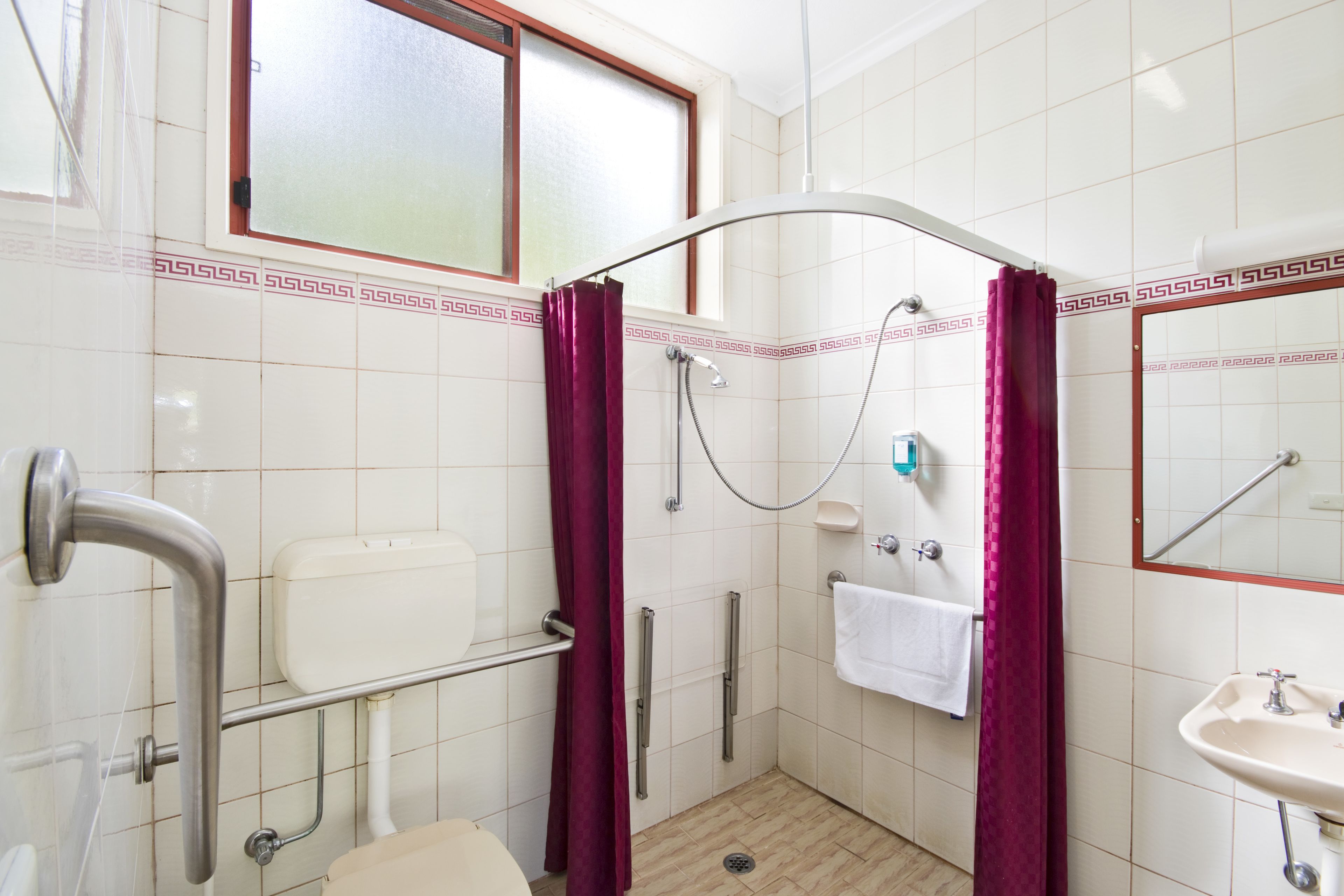 A small, compact bathroom with a shower, toilet, and sink. It features a window, a mirror, and a towel rack.