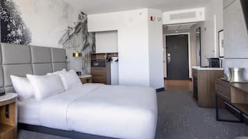 Quarto de hotel moderno com uma cama grande, uma escrivaninha, um guarda-roupa e um banheiro visível através de uma porta aberta.