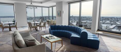 A modern living room with a sectional sofa, a coffee table, and a dining area with chairs and a table. Large windows offer a city view.
