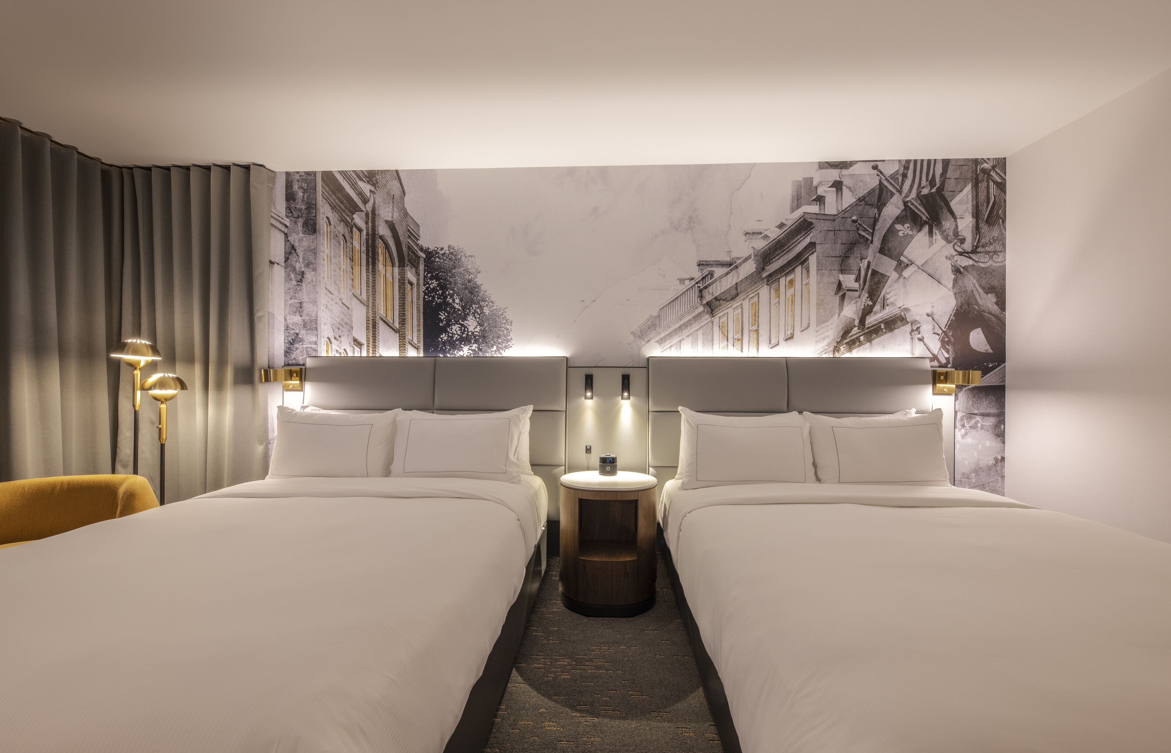 A hotel room with two beds, a nightstand, a lamp, and a mural of a street scene.