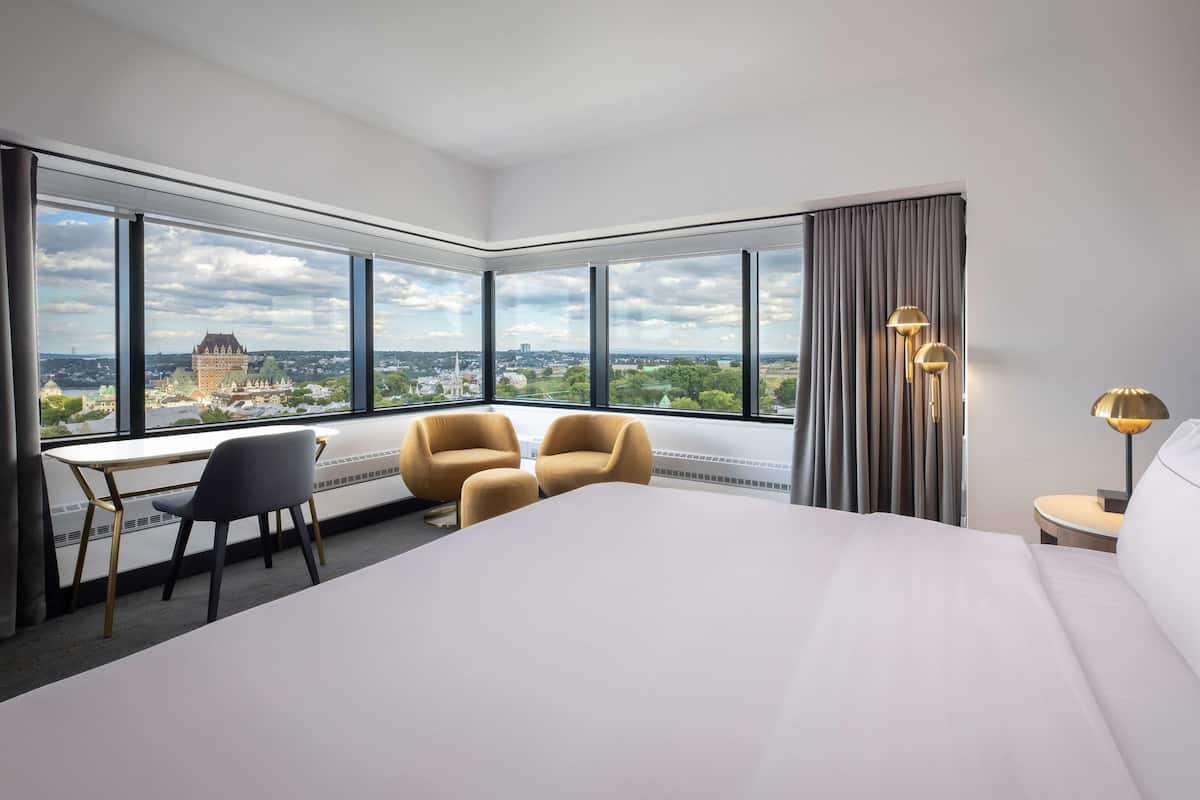 A hotel room with a large bed, two armchairs, a desk, and a view of a cityscape through large windows.