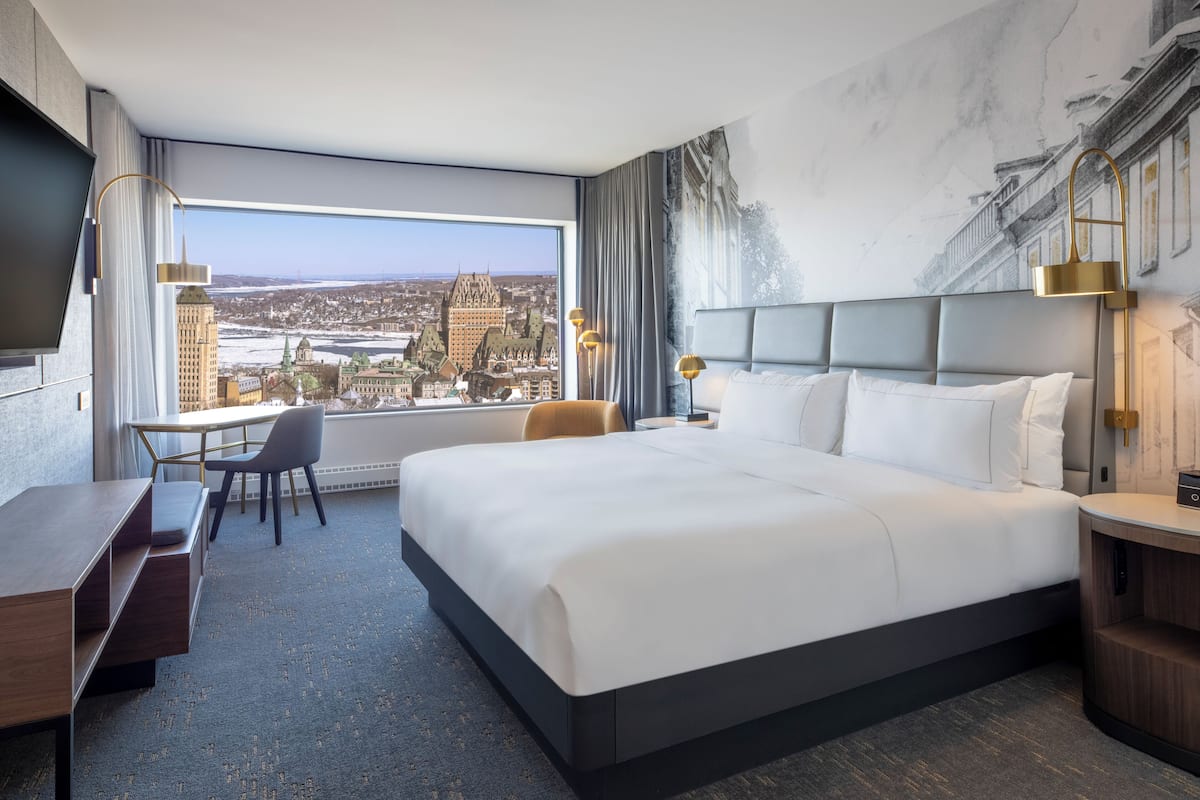 A hotel room with a large bed, a desk, and a chair. There is a view of a cityscape through the window.