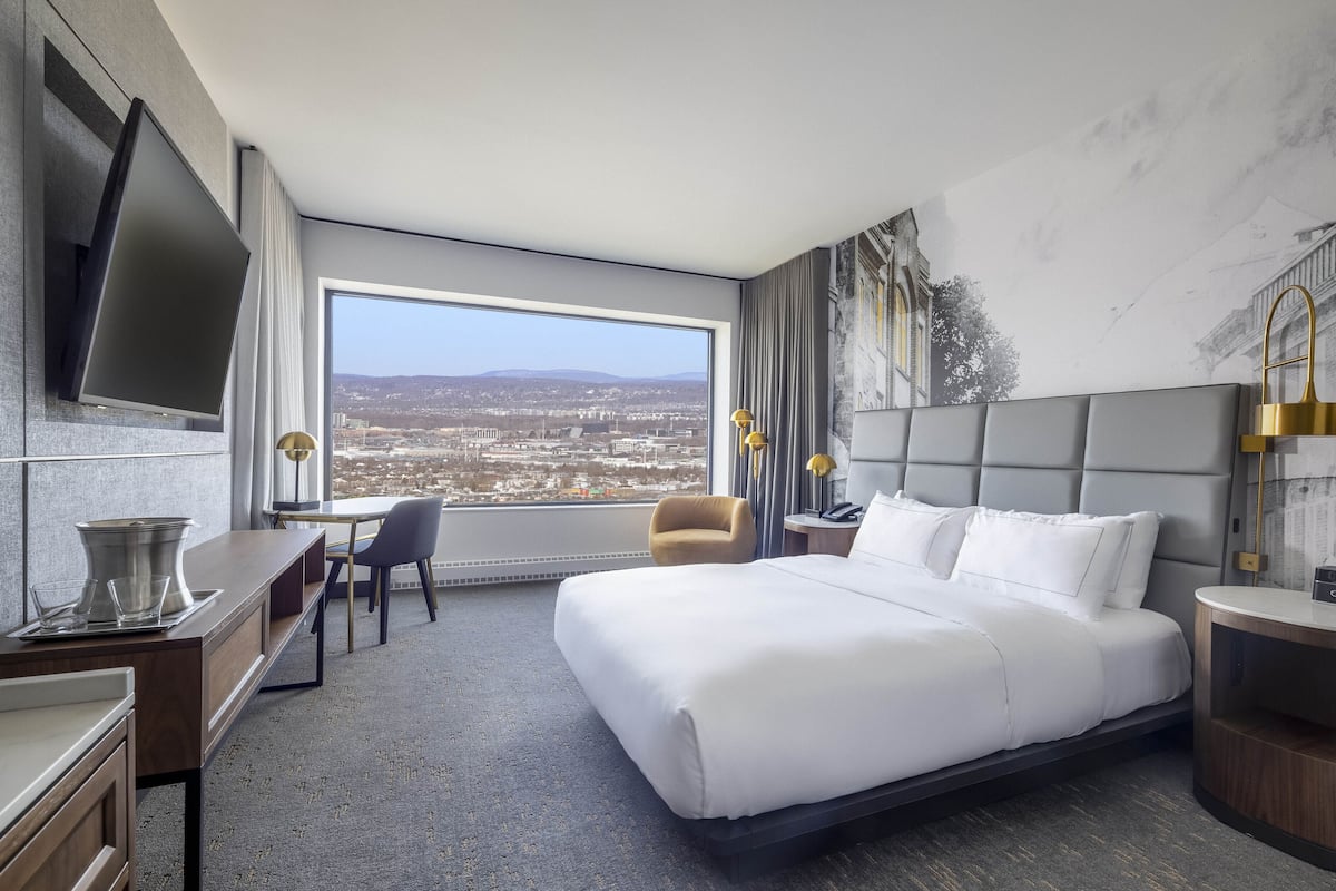 A hotel room with a large bed, a desk, a chair, a TV mounted on the wall, and a window with a city view.