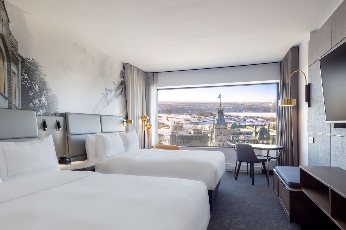 A hotel room with two beds, a desk, and a large window offering a city view.