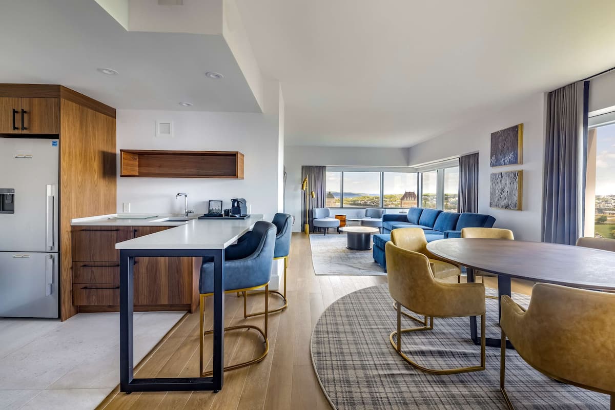 A modern hotel room with a kitchen, dining area, and a living room with a large window offering a city view.