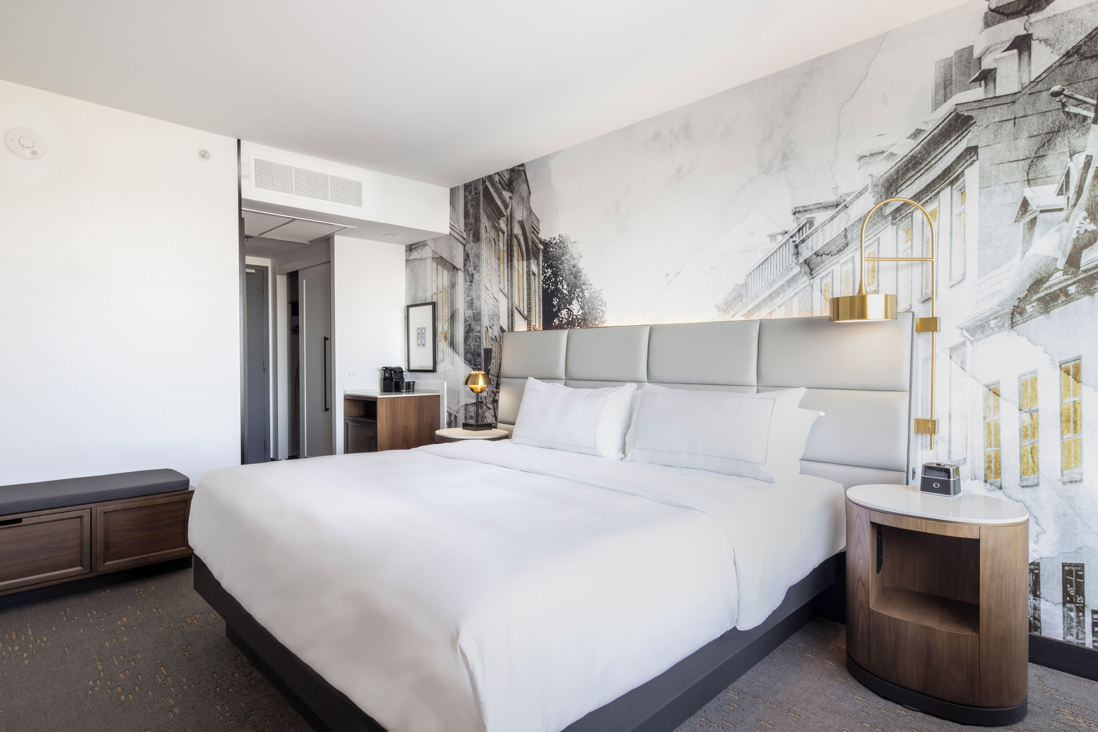 Una habitación de hotel moderna con una cama grande, una mesita de noche con lámpara y un mural en la pared con edificios.