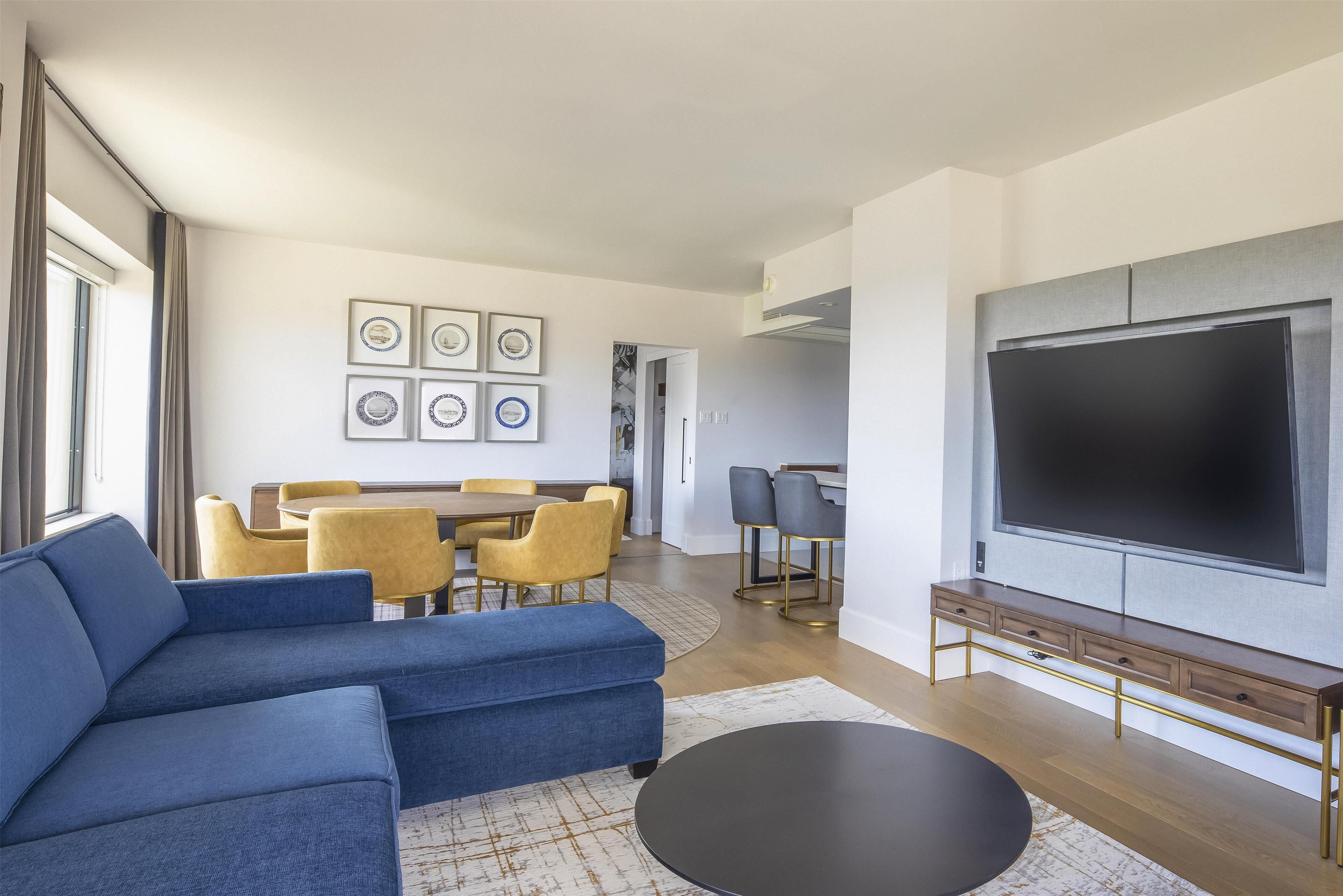 A modern living room with a sectional sofa, a flat-screen TV, and a dining area with yellow chairs.