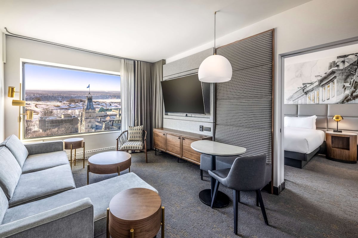 A modern hotel room with a large window offering a city view, a flat-screen TV, a sofa, a dining table, and a bed.