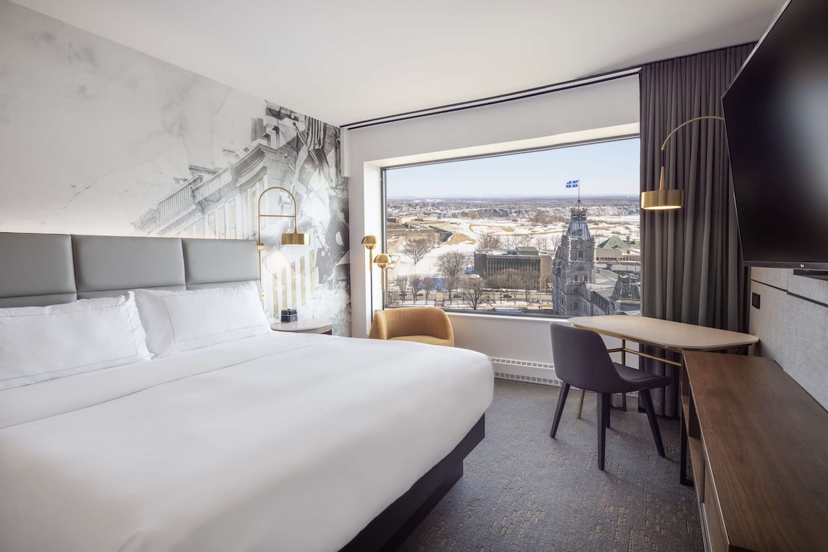 A hotel room with a large bed, a desk, a chair, and a view of a cityscape through a large window.