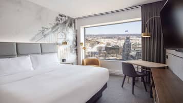 Ett hotellrum med en stor säng, ett skrivbord, en stol och utsikt över stadsbilden genom ett stort fönster.