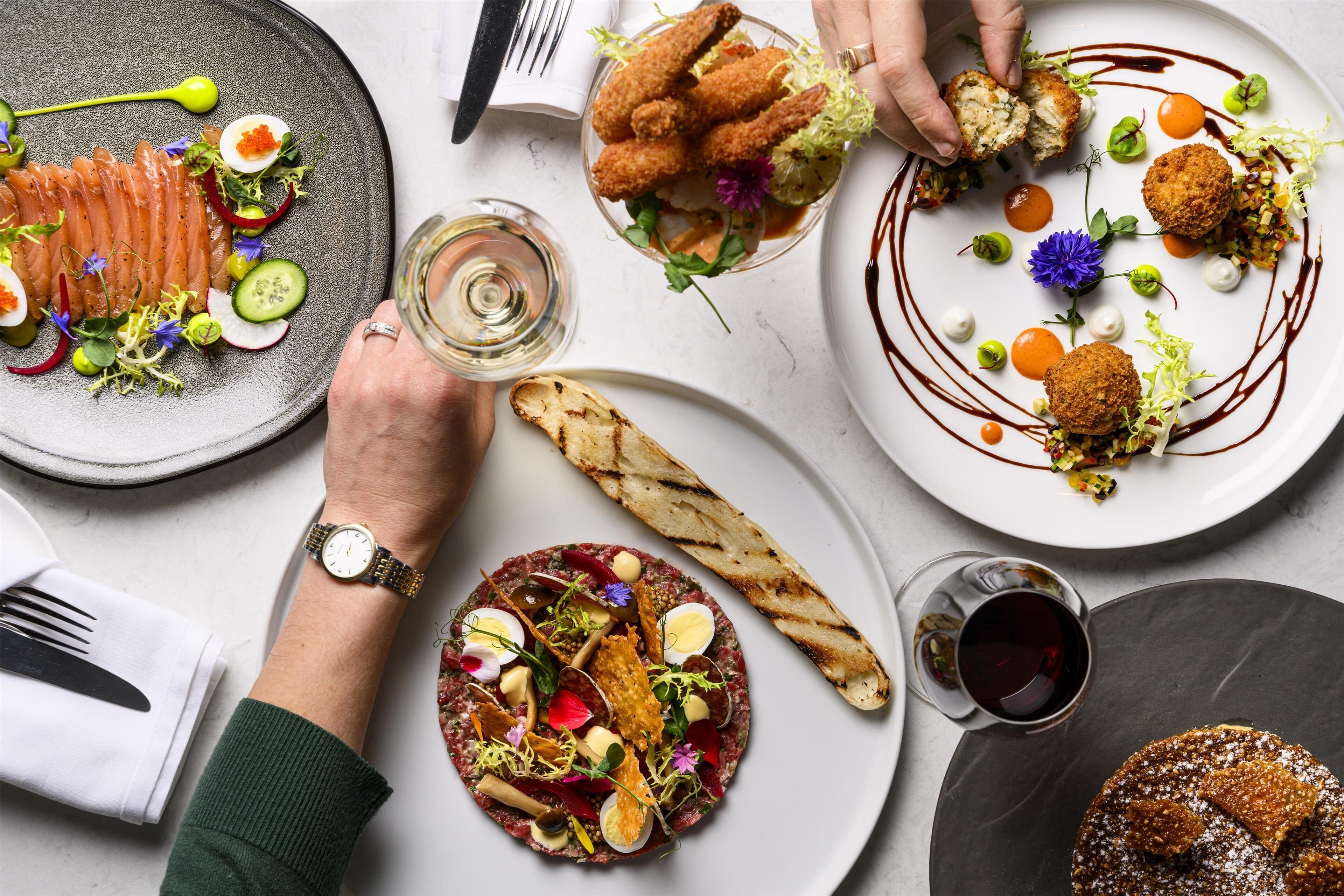 Une variété de plats gastronomiques présentés dans des assiettes, un verre de vin et une personne portant une montre.
