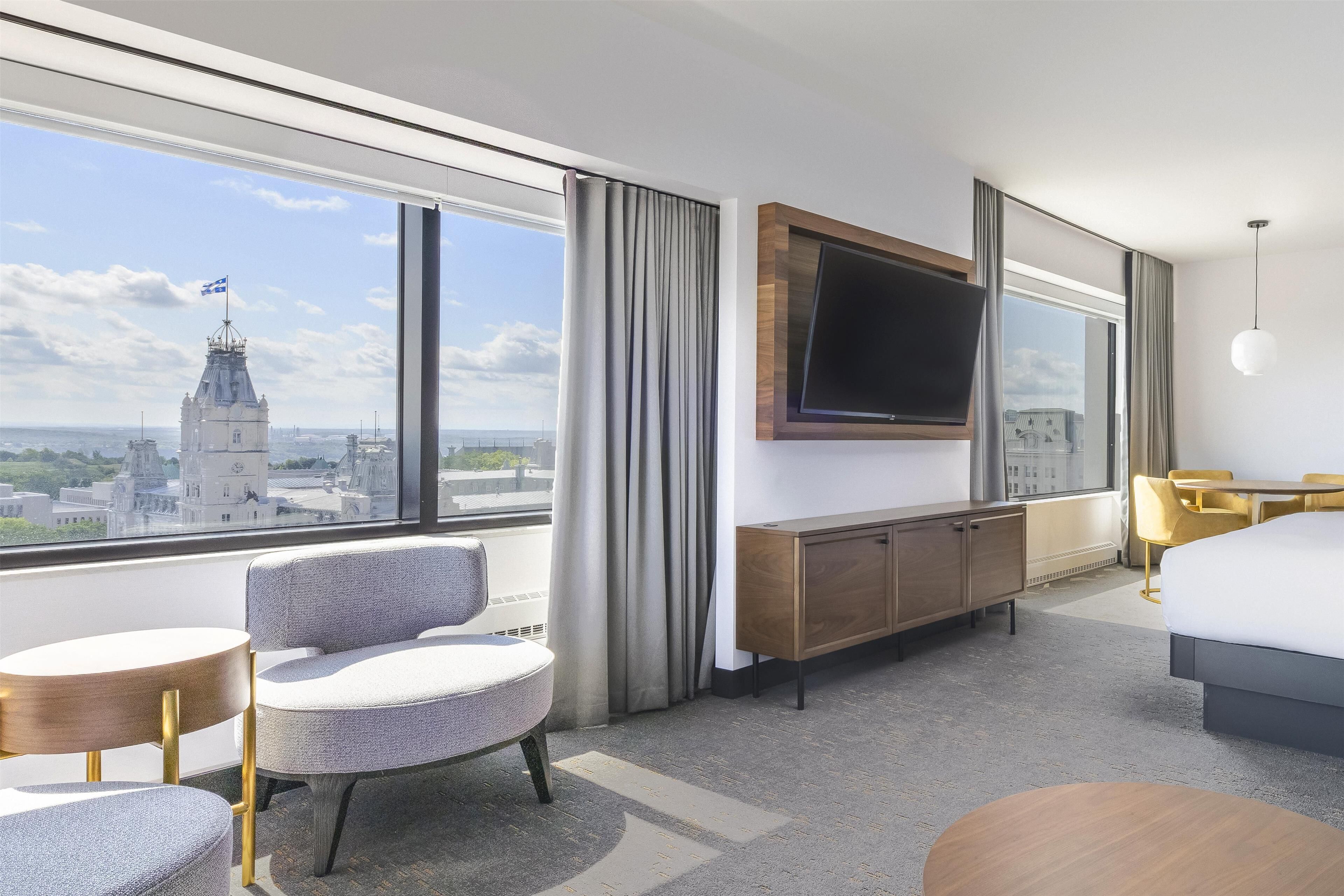 A modern hotel room with a large window, a flat-screen TV, a bed, a desk, and a chair.