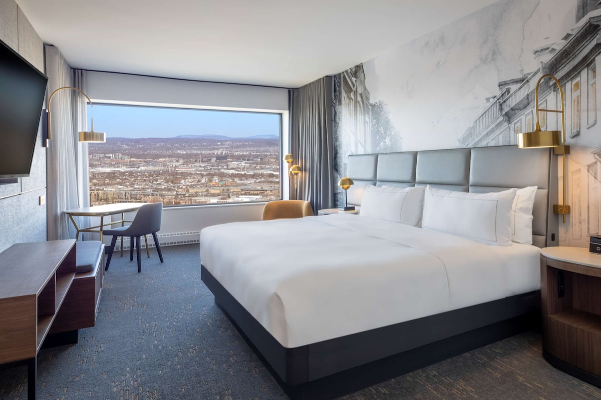A hotel room with a large bed, a desk, and a chair, a view of a cityscape through a large window, and a marble wall with a painting.