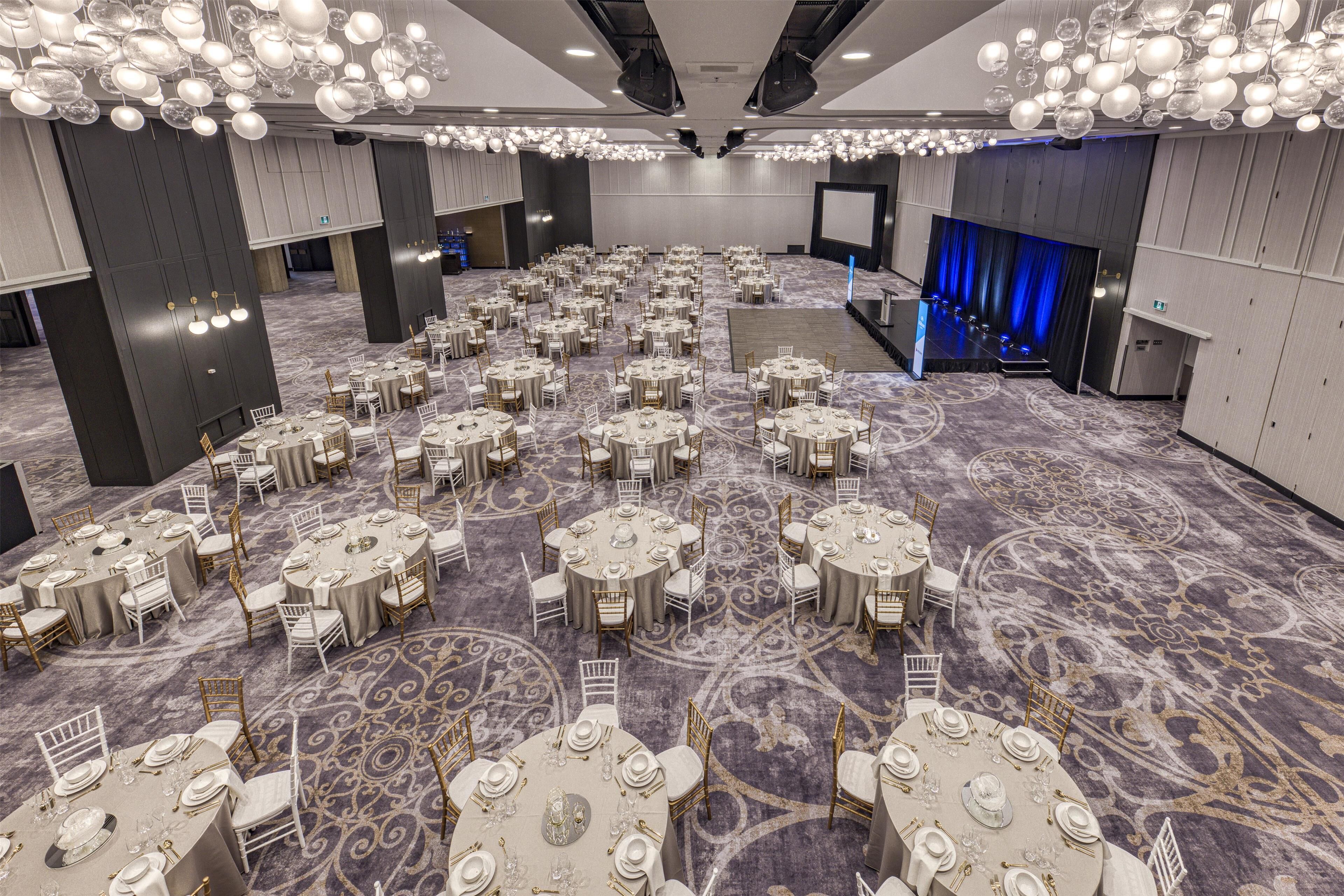A large banquet hall with round tables and chairs, a stage with a blue curtain, and a large screen.