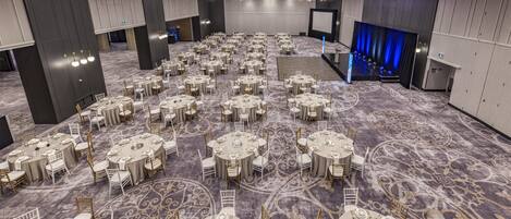 A large banquet hall with round tables and chairs, a stage with a blue curtain, and a large screen.