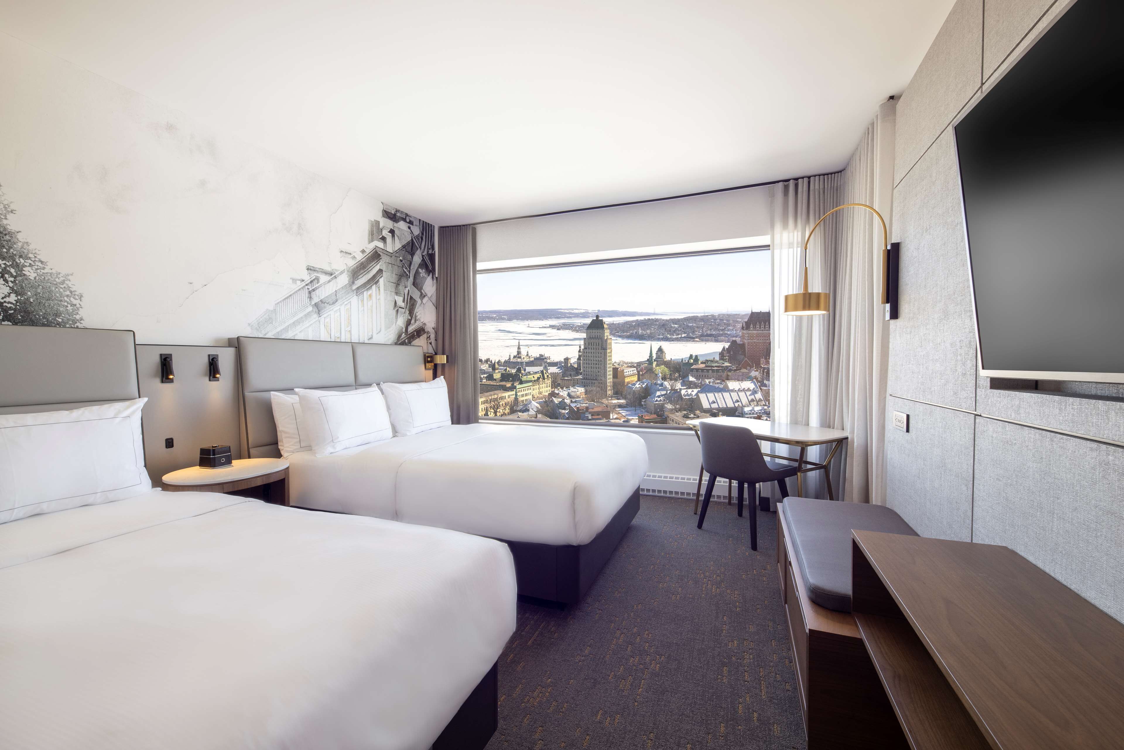 Een hotelkamer met twee bedden, een bureau, een stoel, een televisie en een groot raam met uitzicht op de stad.
