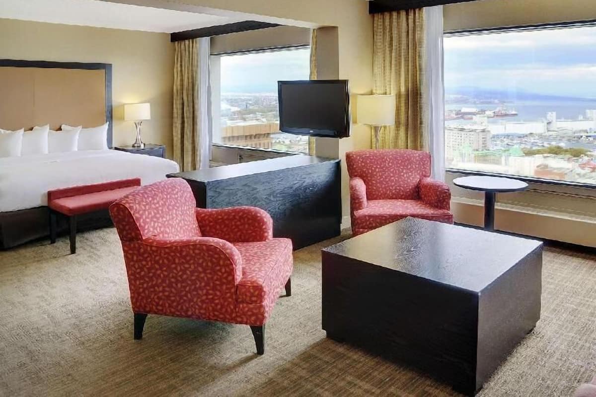 A hotel room with a bed, two armchairs, a coffee table, a TV, and a view of the city.