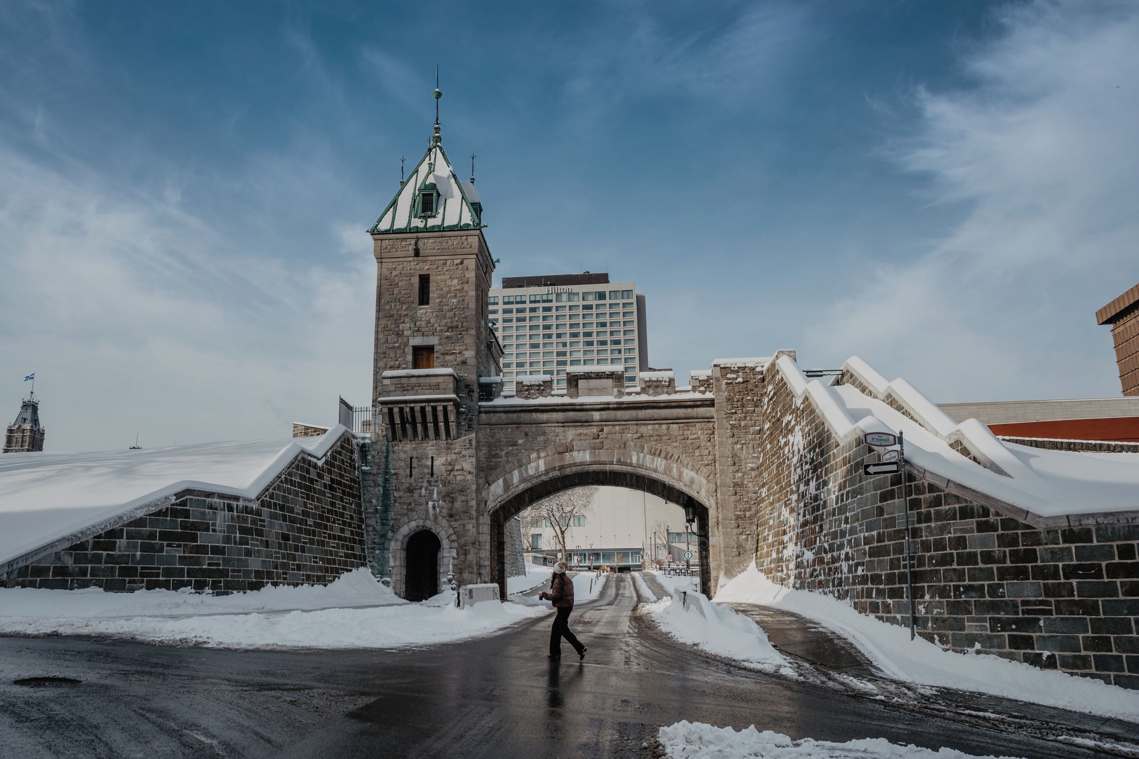 Un pont de pierre historique surmonté d’une tour, une route enneigée et, au loin, un bâtiment moderne.