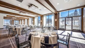 Une vaste salle de banquet avec de nombreuses tables rondes dressées pour le repas, de grandes fenêtres offrant une vue sur l’extérieur et un design de plafond moderne.