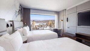 A hotel room with two beds, a large window with a city view, a flat-screen TV, and a small table.