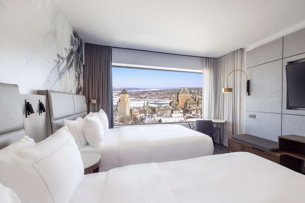A hotel room with two beds, a large window with a city view, a flat-screen TV, and a small table.
