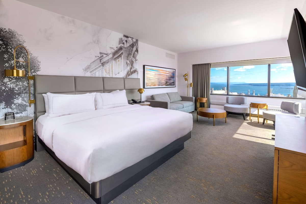 A modern hotel room with a large bed, a sofa, a desk, and a view of the city and sea.