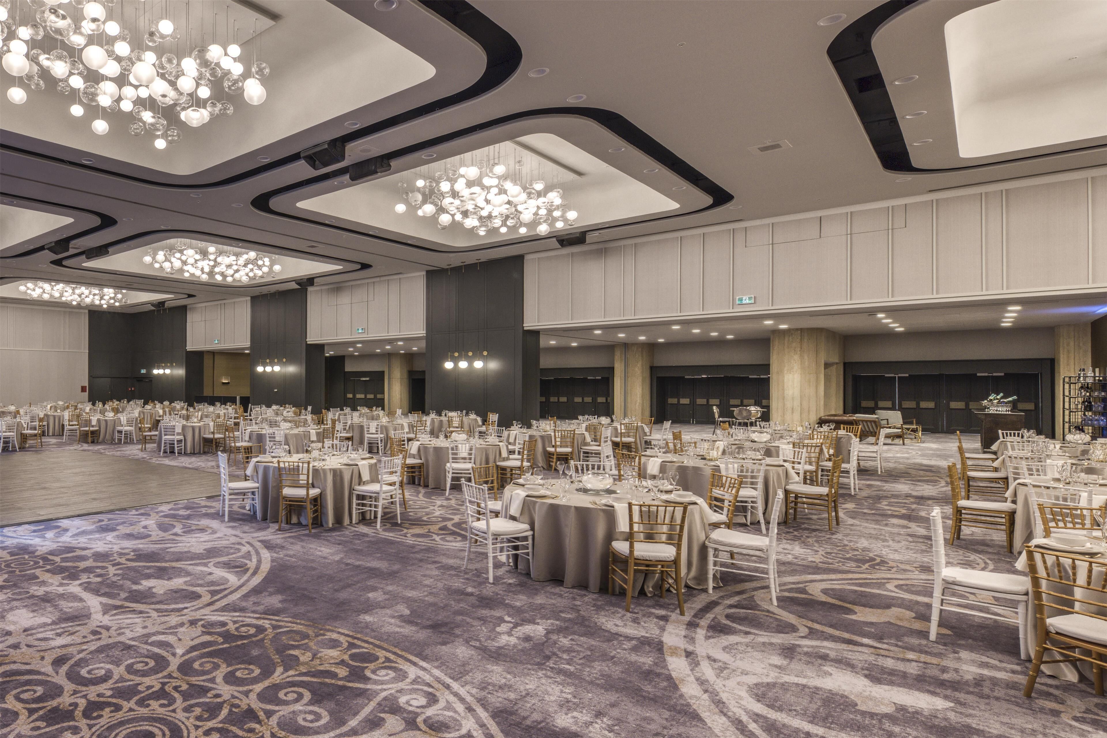 A large banquet hall with round tables and chairs, a patterned carpet, and modern chandeliers.