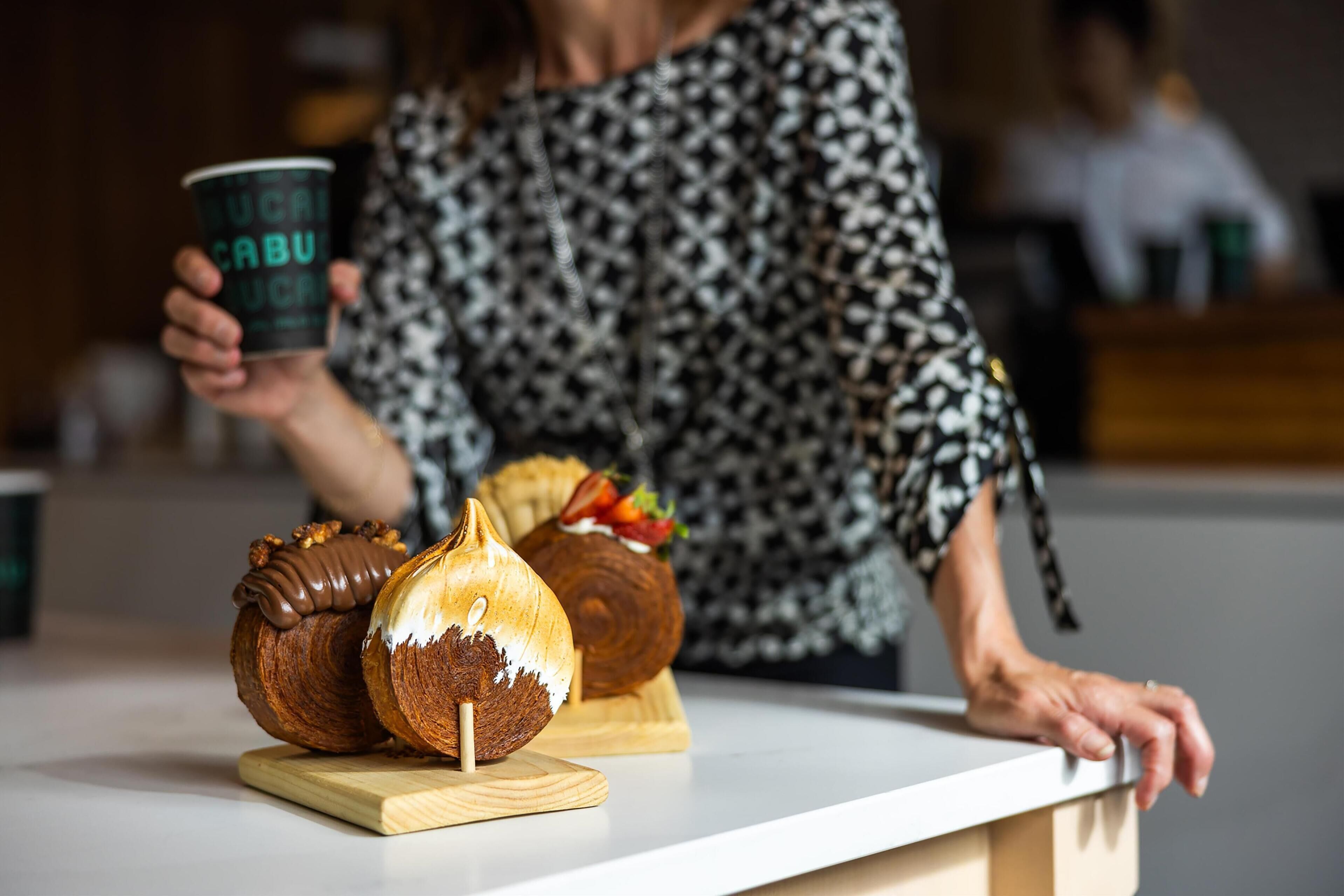 A person holding a cup with text, a loaf of bread, and a pastry on a counter.