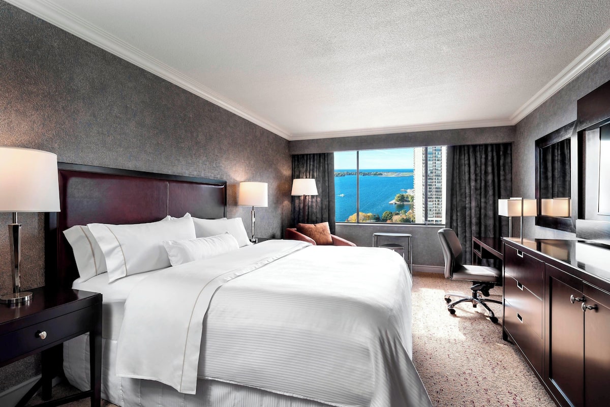 A hotel room with a large bed, two bedside tables with lamps, a desk with a chair, and a view of the ocean.