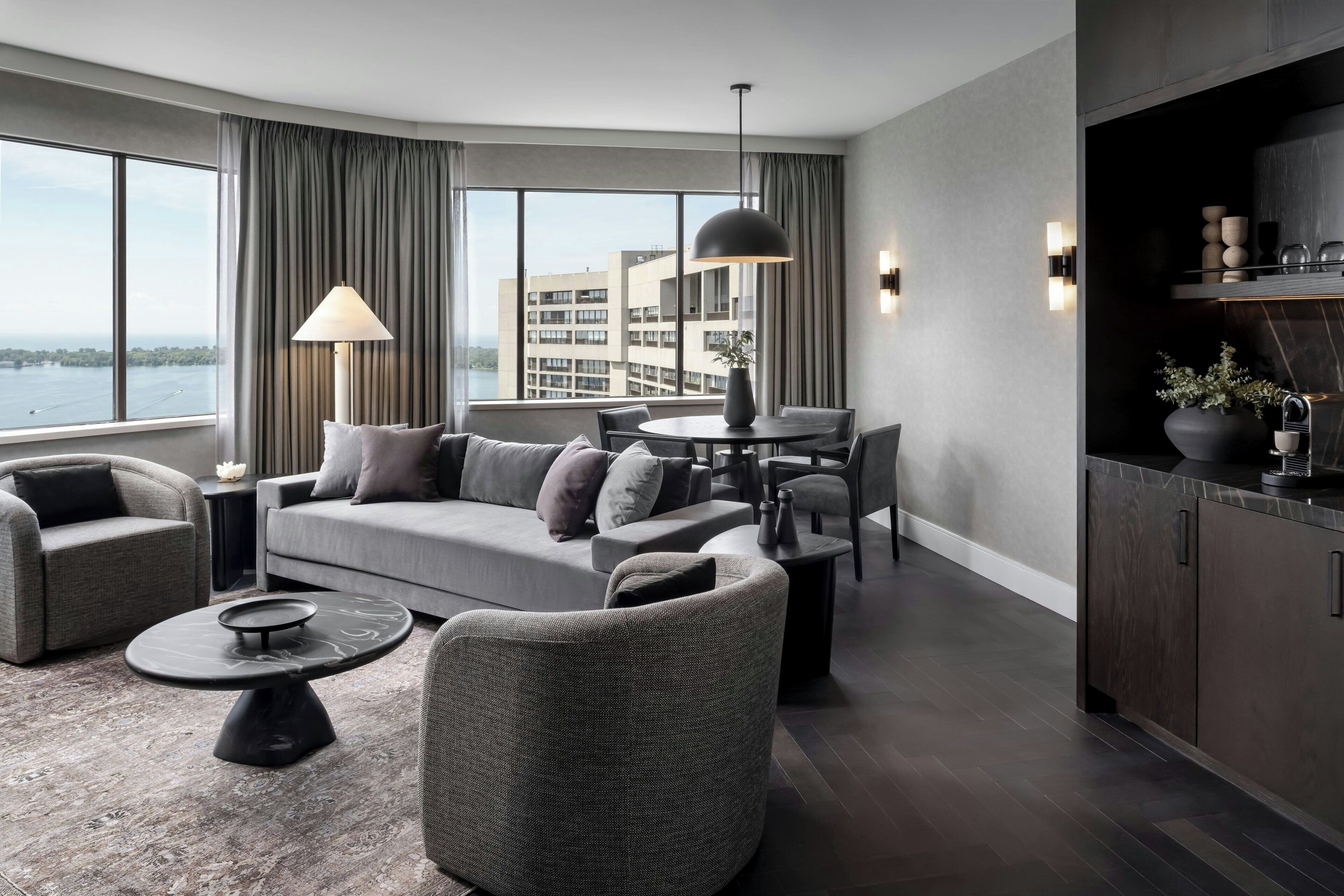 Una sala moderna con un sofá, sillones, una mesa de centro y una mesa de comedor. Hay un ventanal con cortinas, vistas a edificios y una zona de cocina con armarios y una encimera.