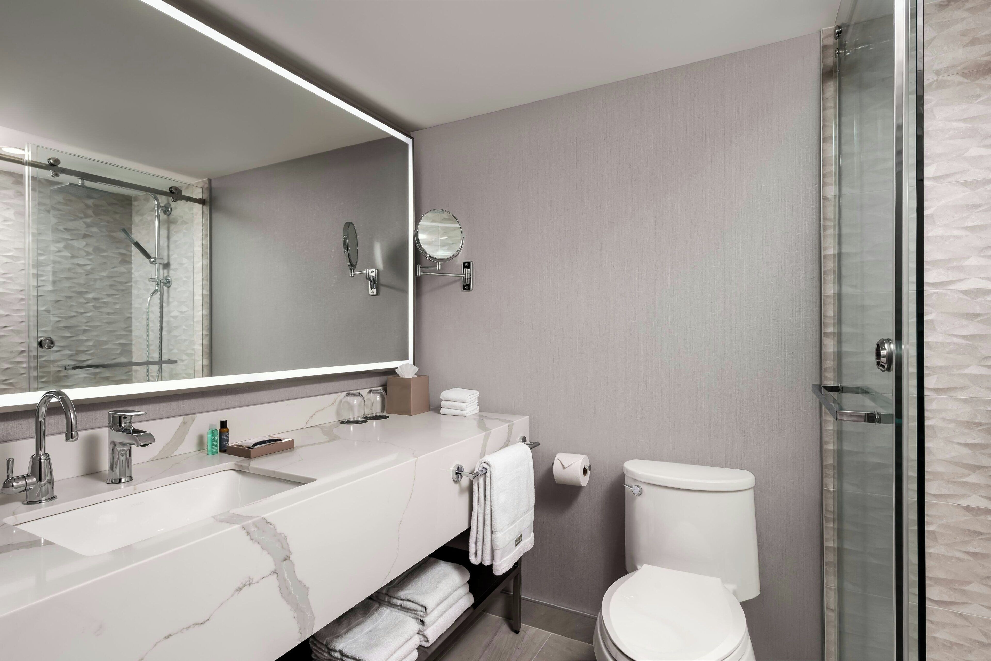 A modern bathroom with a large mirror, a sink with two faucets, a toilet, and a glass-enclosed shower.