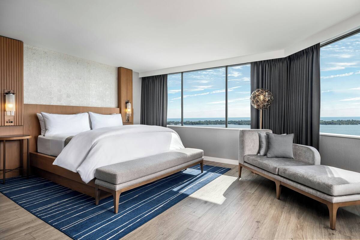 A modern hotel room with a large bed, a sofa, a view of the sea, and a cityscape.