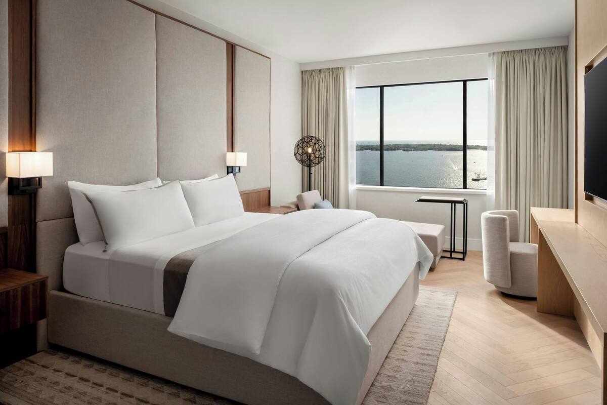 A modern hotel room with a large bed, a desk, a chair, and a view of the water.