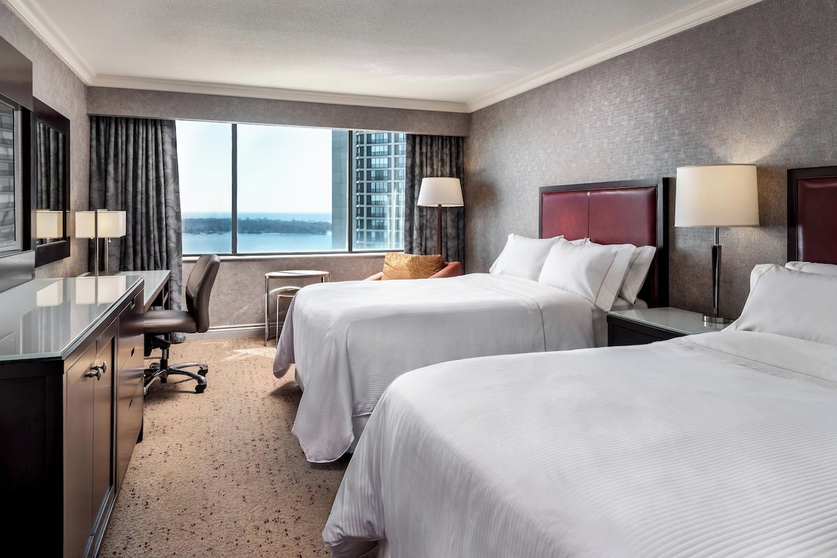 A hotel room with two beds, a desk, a chair, and a large window offering a city view.