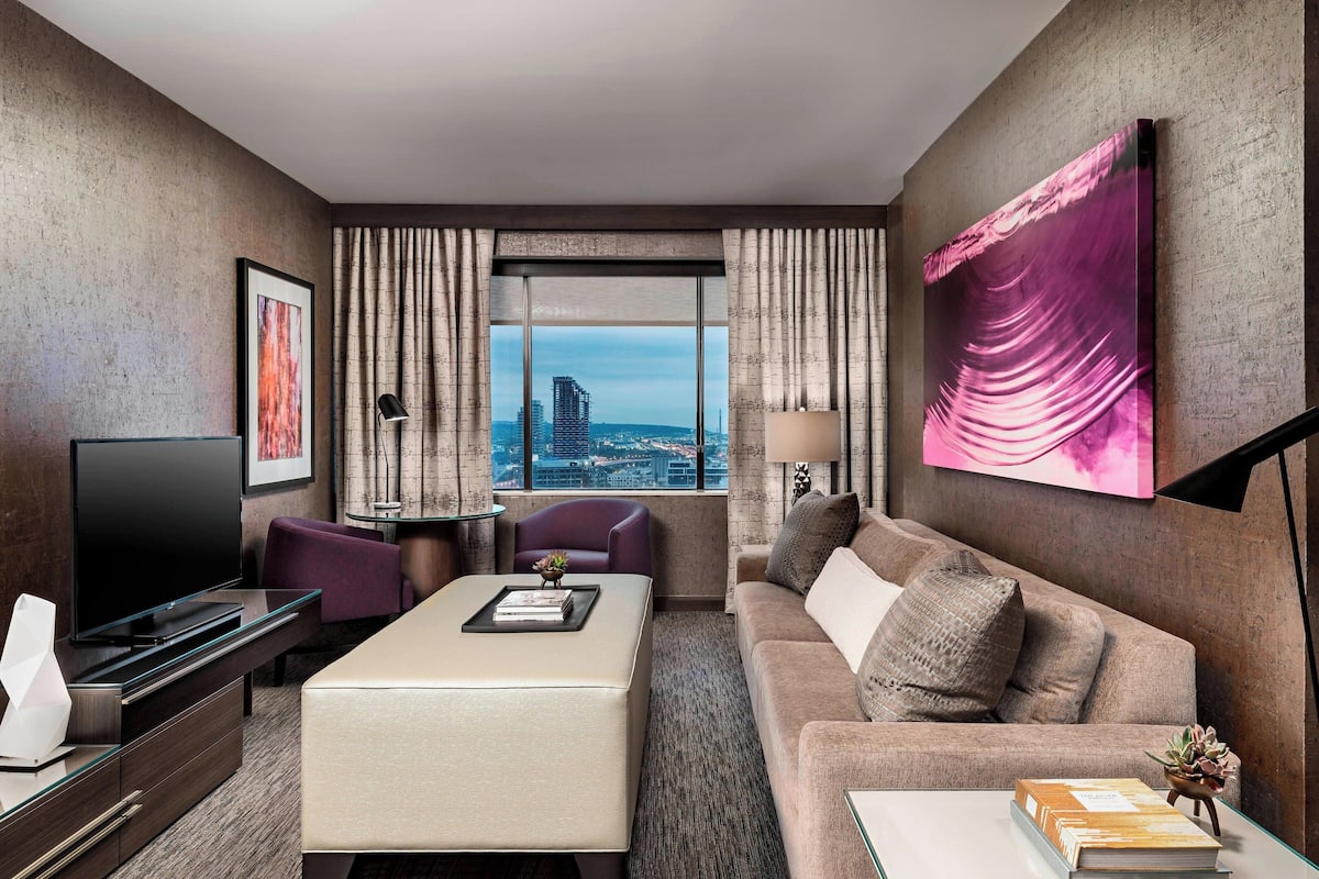 A modern hotel room with a large window, a flat-screen TV, a sofa, a coffee table, and a desk with a lamp.