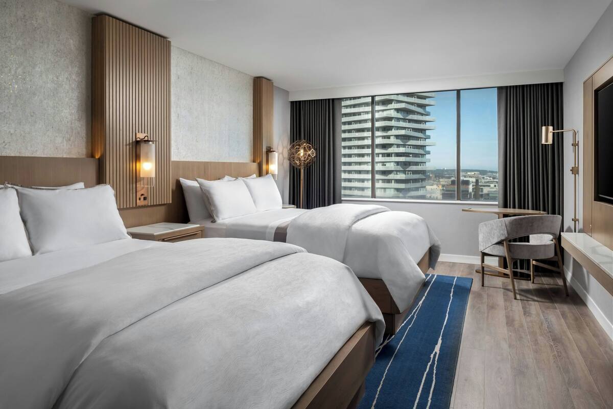 A hotel room with two beds, a desk, a chair, a large window with a city view, and a TV.
