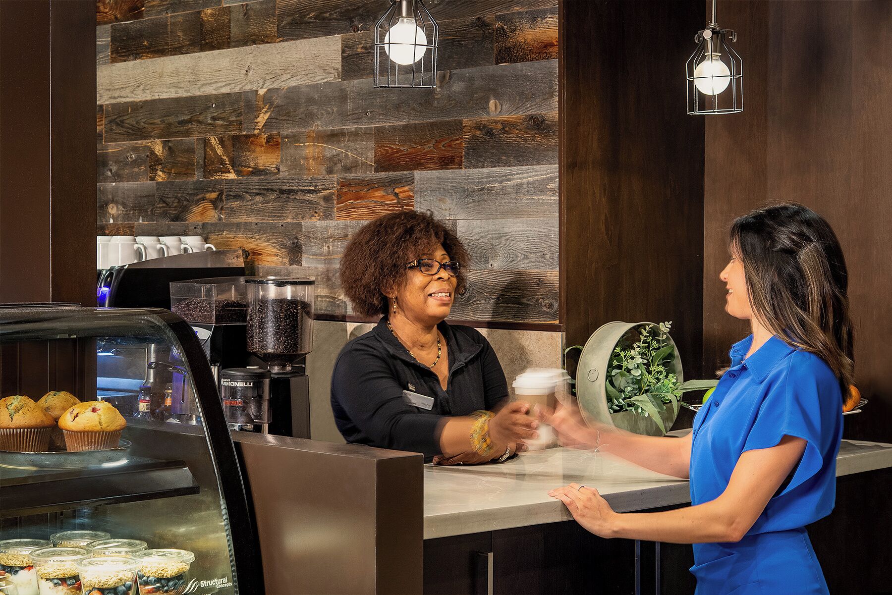 A coffee shop interior with a rustic wooden wall, a coffee machine, muffins, and a counter where a customer and a barista are interacting.