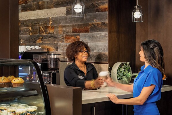 A coffee shop interior with a rustic wooden wall, a coffee machine, muffins, and a counter where a customer and a barista are interacting.