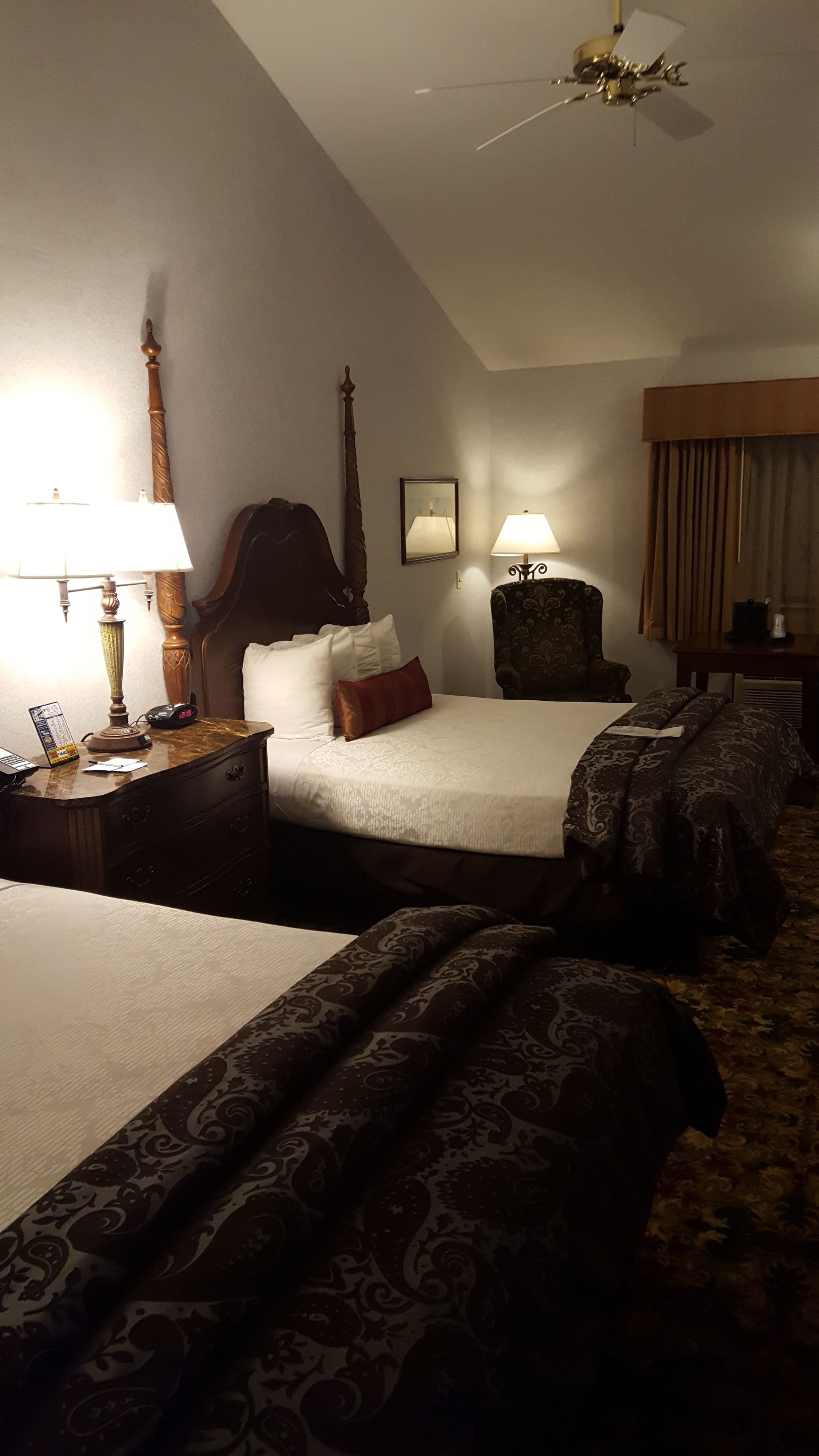 Une chambre d’hôtel avec deux lits, une table de chevet avec une lampe, une chaise, un ventilateur de plafond et une fenêtre avec des rideaux.